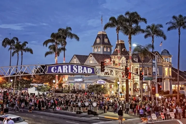 Local crowd enjoying the Carlsbad Village Street Fair at dusk, with historic buildings, palm trees, and the iconic Carlsbad sign.